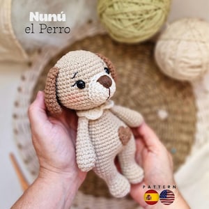 May include: A small, beige crocheted dog toy with brown ears, a dark brown nose, and black eyes. The toy features a small brown heart patch on its chest and a white collar. The text 'Nunú el Perro' is visible in the upper left corner of the image.