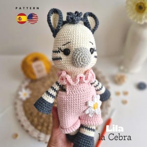 Puede incluir: Muñeca cebra de ganchillo hecha a mano con un atuendo rosa y una margarita blanca. La cebra tiene brazos y piernas a rayas blancas y negras, y una nariz gris. El texto "Lila la Cebra" es visible.