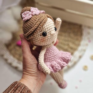 Op de afbeelding: Handgemaakte gehaakte pop in de vorm van een ballerina. De pop heeft lichtbruin haar, een roze strik en een roze tutu. De armen van de pop zijn omhoog, en ze heeft zwarte ogen en roze wangen. De pop is ongeveer 15 cm hoog.