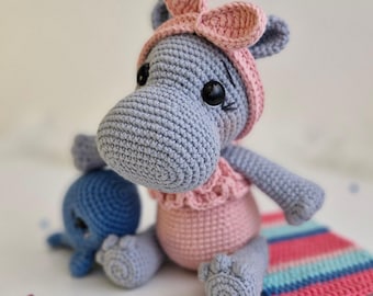 May include: A gray crocheted hippopotamus wearing a pink crocheted dress and headband. The hippopotamus is sitting on a colorful striped blanket with a smaller blue crocheted octopus beside it.