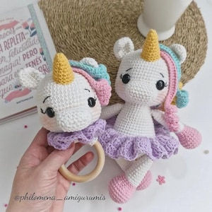 Puede incluir: Dos juguetes de unicornio hechos a mano con ganchillo. Uno es un sonajero con un anillo de madera, el otro es una muñeca. Ambos tienen cuerpos blancos, cuernos amarillos y detalles rosas, azules y morados. La muñeca tiene un tutú lavanda y pies rosas.