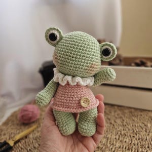 Puede incluir: Una muñeca de rana de ganchillo hecha a mano en verde claro y rosa. La rana tiene ojos negros grandes, un cuello con volantes blancos y un vestido rosa con un botón de madera. La muñeca mide aproximadamente 13 cm de alto.