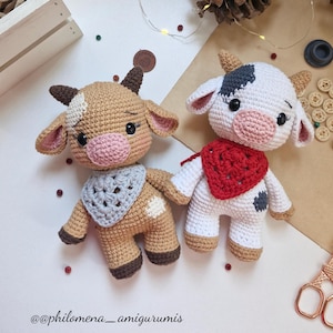 Puede incluir: Dos figuras de vaca de ganchillo hechas a mano. Una es de color canela con un pañuelo azul, la otra es blanca con un pañuelo rojo. Ambas tienen narices rosas, ojos negros y cuernos pequeños. Las figuras están sobre un fondo neutro.