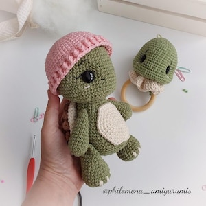 Puede incluir: Una tortuga de crochet verde con un gorro de crochet rosa y un sonajero de tortuga de crochet verde más pequeño con un anillo de madera. La tortuga más grande tiene ojos negros y un caparazón blanco.