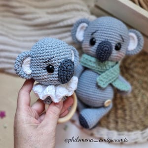 Puede incluir: Dos juguetes de koala hechos a mano con ganchillo. Uno es un sonajero con un anillo de madera y un cuello con volantes blancos. El otro es un koala más grande con una bufanda verde. Ambos son grises con ojos y narices negros.