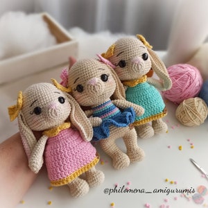 May include: Three crocheted bunny dolls wearing dresses. The bunnies are beige with black eyes and yellow bows. The dresses are pink, blue and turquoise.