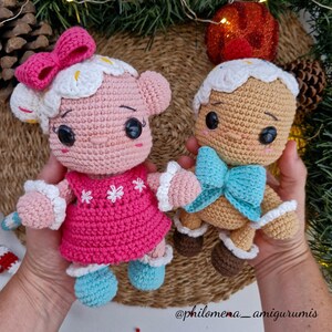Puede incluir: Dos muñecas de jengibre de crochet. Una es rosa con un vestido blanco y rosa y un lazo azul. La otra es marrón con un vestido blanco y marrón y un lazo azul. Ambas muñecas tienen ojos negros y mejillas rojas.