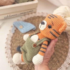 Puede incluir: Amigurumi de tigre tejido a ganchillo, naranja con rayas negras, con un peto verde con botones de madera. El tigre tiene patas blancas y una cara blanca con ojos negros. El texto "@philomena_amigurumis" es visible.