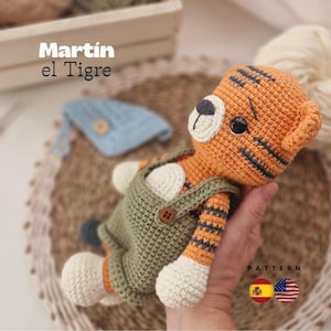 Puede incluir: Juguete de tigre tejido a ganchillo en naranja y negro, con un peto verde. El juguete tiene detalles blancos en las patas y la cara. El texto "Martín el Tigre" es visible. Un pequeño sombrero azul está en el fondo.