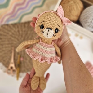 Puede incluir: Una muñeca de foca de ganchillo hecha a mano en color beige, con un top a rayas rosas y blancas y un lazo rosa a juego. La foca tiene ojos negros y un hocico color crema. La muñeca está siendo sostenida en las manos.