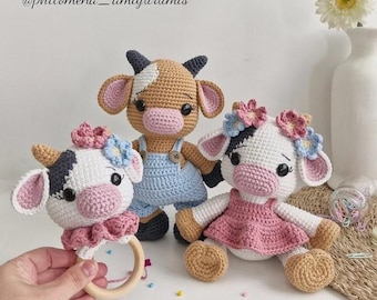 Puede incluir: Tres muñecas de vaca de ganchillo. Una es un sonajero con un anillo de madera, y las otras dos son peluches. Las vacas tienen patrones en blanco y negro o marrón y blanco, orejas rosas y coronas de flores. Una vaca lleva un vestido rosa y otra un peto azul.