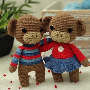 Puede incluir: Dos juguetes de mono de crochet, uno con un suéter a rayas azul y rojo y el otro con una camisa roja y una falda azul.