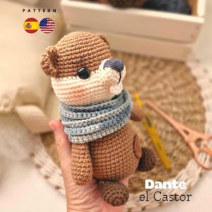 May include: A handmade crochet otter toy in shades of brown, white, and blue. The otter has a blue and white striped scarf and black button eyes. The text "Dante el Castor" is visible.