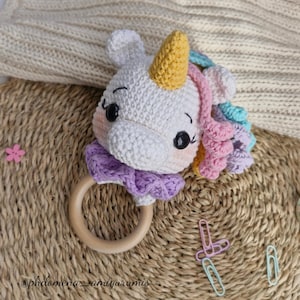 Puede incluir: Un sonajero de unicornio de ganchillo hecho a mano, con cuerpo blanco, cuerno amarillo y melena colorida. El sonajero tiene un anillo de madera y un cuello morado. El texto "@philomena_amigurumis" es visible en la parte inferior.