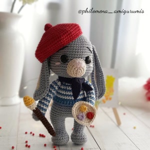 Puede incluir: Un conejito de crochet gris que lleva una boina roja, una camisa a rayas azul y blanca y una bufanda azul. El conejito sostiene un pincel y una paleta con pintura de colores.