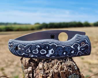 May include: A black and white patterned pocket knife with a silver blade and a round hole in the handle. The knife is sitting on a weathered wooden post.