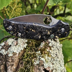 May include: A folding pocket knife with a silver blade and a black and gold handle. The handle has a textured carbon fiber appearance with gold flecks. The knife is resting on a moss-covered wooden surface, with green foliage in the background.