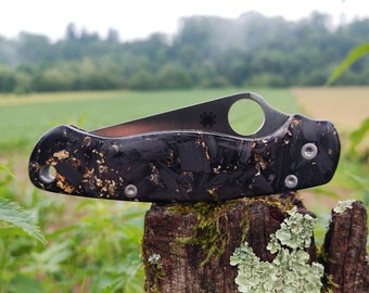 May include: A black and gold pocket knife with a textured handle sits on a weathered wooden post. The knife has a small hole in the blade for opening it with one hand.