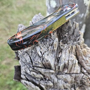 May include: A folding pocket knife with a black and red patterned handle. The blade is partially visible, and the knife is resting on a weathered wooden surface. The handle features a glossy finish with intricate red swirls.