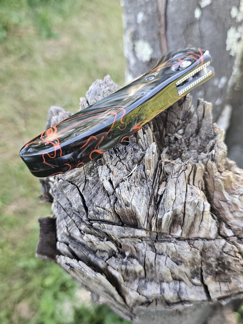 May include: A folding pocket knife with a black and red patterned handle. The blade is partially visible, and the knife is resting on a weathered wooden surface. The handle features a glossy finish with intricate red swirls.