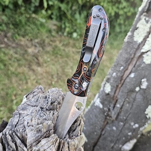 May include: A Benchmade folding knife with a partially serrated blade is embedded in a piece of weathered wood. The knife features a black handle with orange and brown patterns and a silver pocket clip with the word "BENCHMADE".
