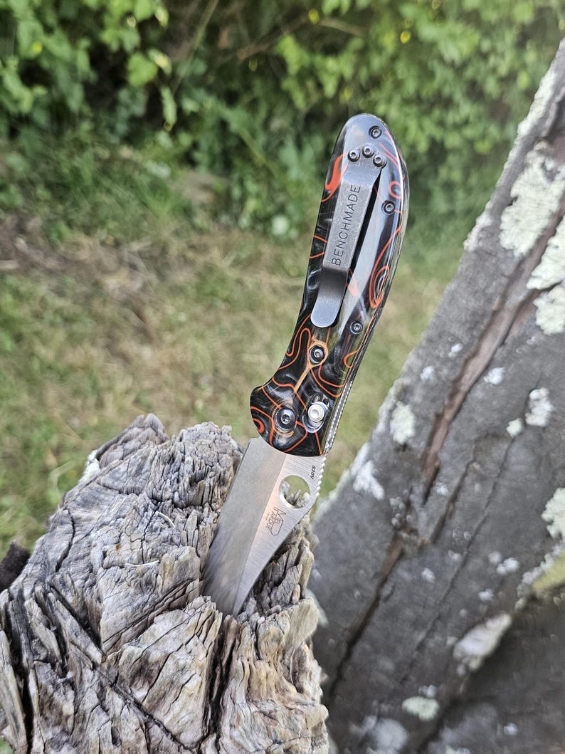 May include: A Benchmade folding knife with a partially serrated blade is embedded in a piece of weathered wood. The knife features a black handle with orange and brown patterns and a silver pocket clip with the word "BENCHMADE".