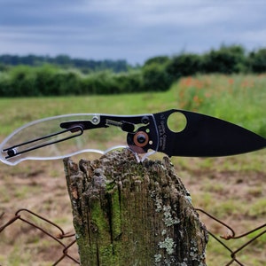 Puede incluir: Un cuchillo de bolsillo negro con un mango de plástico transparente. El cuchillo está sentado en un poste de cerca de madera con una cerca de alambre en el fondo.