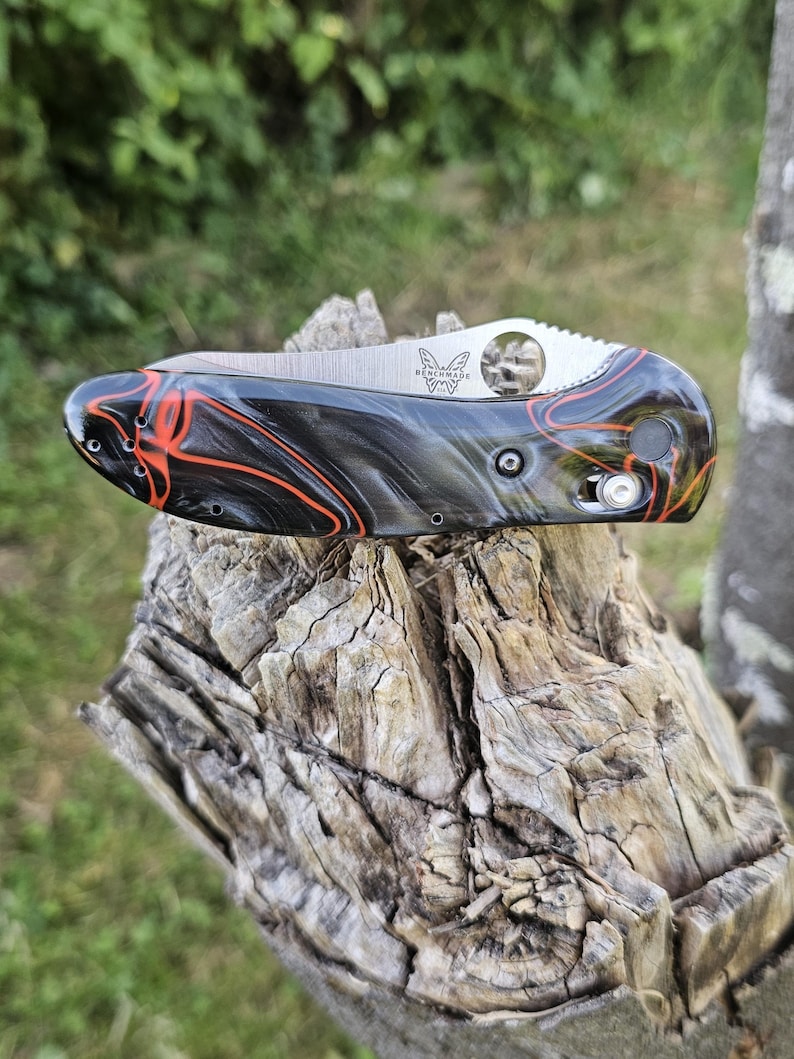 May include: A folding knife with a stainless steel blade and a handle with a black and red marbled design. The knife is on a weathered wooden surface, with a blurred green background.