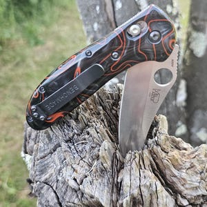 May include: A Benchmade folding knife with a partially serrated blade. The handle is black with an orange and red pattern. The knife has a black pocket clip with the word "BENCHMADE" printed on it. The blade is silver.