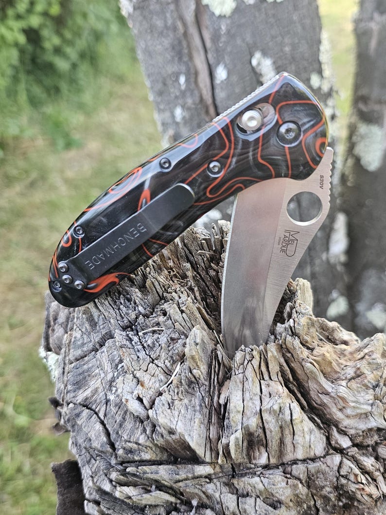 May include: A Benchmade folding knife with a partially serrated blade. The handle is black with an orange and red pattern. The knife has a black pocket clip with the word "BENCHMADE" printed on it. The blade is silver.