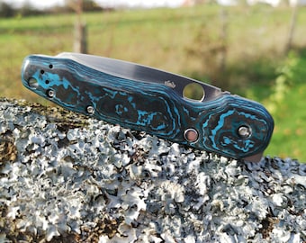 May include: A blue and black pocket knife with a textured handle. The knife is closed and resting on a piece of bark.