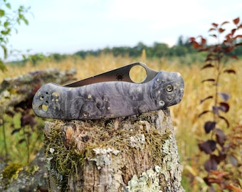 May include: A black and grey pocket knife with a wood grain pattern and a silver blade. The knife is sitting on a tree stump with moss growing on it.