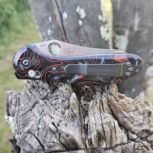 May include: A Benchmade folding knife with a black and orange marbled handle. The blade is silver with the text "MEL LARUE" and "S30V". The pocket clip is silver and has the word "BENCHMADE" printed on it.