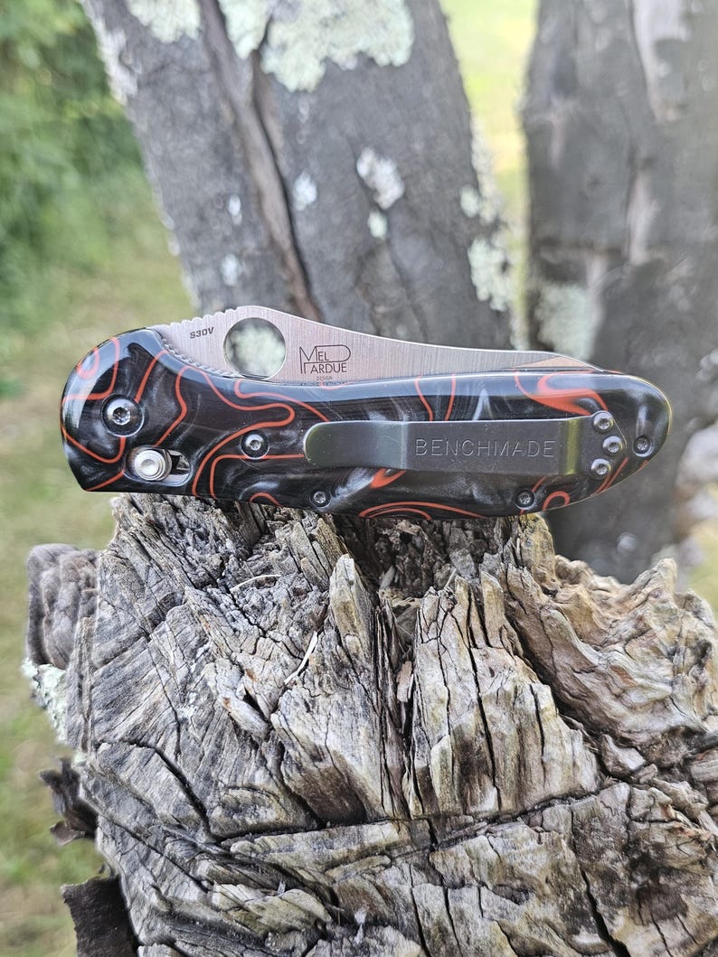 May include: A Benchmade folding knife with a black and orange marbled handle. The blade is silver with the text "MEL LARUE" and "S30V". The pocket clip is silver and has the word "BENCHMADE" printed on it.
