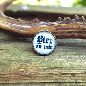 Könnte beinhalten: Ein runder Metallknopf mit dem deutschen Text "Bier zu mir" in blauer Schrift auf weißem Hintergrund. Der Knopf hat einen bronzefarbenen Rand und liegt auf einer Holzoberfläche.