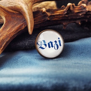 Könnte beinhalten: Ein runder, messingfarbener Ansteckpin mit dem Wort "Bazi" in blauer gotischer Schrift auf weißem Hintergrund. Der Pin befindet sich vor einem blauen Stoff, einem dunkelgrauen Stoff und einem braunen Geweih.