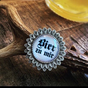 Könnte beinhalten: Eine silberne Anstecknadel mit weißem Hintergrund und schwarzem Text, der "Bier zu mir" lautet. Die Anstecknadel ist blumenförmig mit einem Kreis in der Mitte.