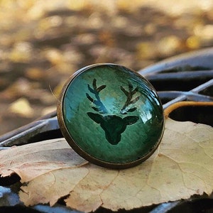 Könnte beinhalten: Ein grüner Glascabochon mit einer bronzenen Fassung zeigt eine Silhouette eines Hirsches mit Geweih. Der Cabochon liegt auf einem braunen Blatt.