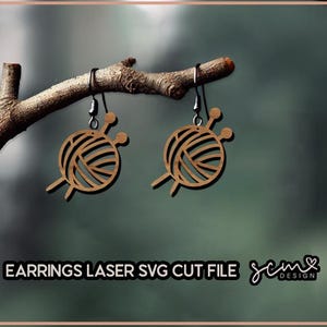 May include: A pair of wooden earrings shaped like yarn balls with knitting needles. The earrings are hanging from a branch. The text "EARRINGS LASER SVG CUT FILE jcm DESIGN" is visible at the bottom of the image.