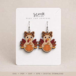 May include: A pair of wooden dangle earrings featuring a cartoon turkey holding a pumpkin, wearing a leopard print bow. The turkey has brown, red, and orange feathers. The display card features the text SCM DESIGN MADE FOR SOMEONE.