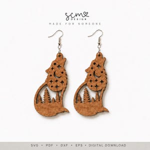 May include: A pair of brown wooden earrings featuring a laser-cut design of a howling wolf silhouette. The wolf body contains cutouts of a crescent moon, stars, and a forest of pine trees. The earrings hang from silver-toned metal fishhook wires.