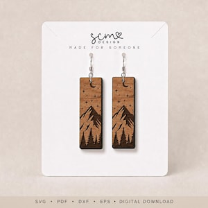 May include: A pair of rectangular wooden drop earrings featuring a laser-cut mountain range, pine trees, stars, and a crescent moon. The earrings are displayed on a white card with the text SCM DESIGN MADE FOR SOMEONE and SVG PDF DXF EPS DIGITAL DOWNLOAD.