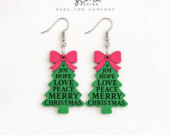 May include: A pair of dangle earrings shaped like green Christmas trees with a pink bow at the top. The trees feature the black text: JOY, HOPE, LOVE, PEACE, MERRY CHRISTMAS. The earrings hang from silver-toned metal hooks and are displayed with the text SCM DESIGN MADE FOR SOMEONE.