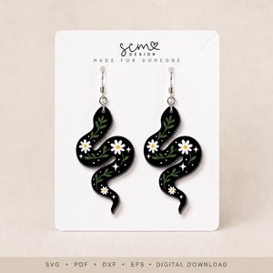 May include: A pair of black snake-shaped dangle earrings featuring white daisy flowers, green leafy vines, and small white star accents. The earrings are displayed on a white card with the text scm DESIGN MADE FOR SOMEONE at the top.