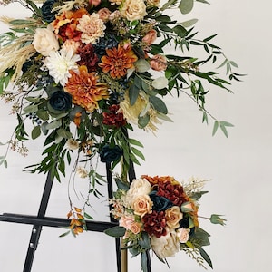 Può includere: Un arco da matrimonio realizzato con fiori artificiali in tonalità arancio, pesca e crema, con verde e erbe secche. L'arco è esposto su un cavalletto nero.