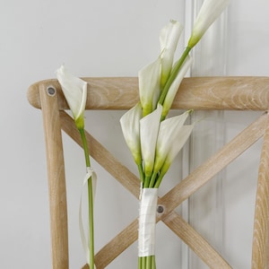 Op de afbeelding: Een boeket witte calla lelies met groene stelen, samengebonden met een wit satijnen lint. Een enkele bijpassende calla lelie staat apart aan de linkerkant, ook vastgebonden met een dun wit lint, tegen een lichte houten stoel met kruisrug.