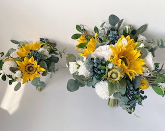 Pode incluir: Dois buquês de flores artificiais, um grande e outro pequeno, com girassóis amarelos, flores brancas e bagas azuis. Os buquês são organizados com folhagem.