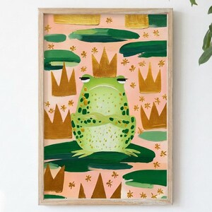 May include: A framed art print featuring a green frog wearing a gold crown, with arms crossed. The frog is on a lily pad, surrounded by gold crowns, green lily pads, and small flowers, all set against a pink background. The artwork has a whimsical, illustrative style.