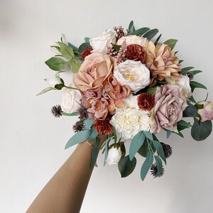 May include: A bouquet of blush, cream, and burgundy artificial flowers with greenery. The bouquet is held by a hand.