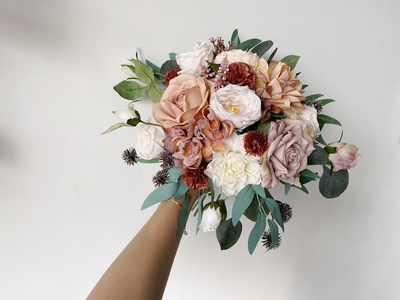 May include: A bouquet of blush, cream, and burgundy artificial flowers with greenery. The bouquet is held by a hand.